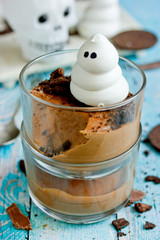 Halloween dessert for kids - delicious chocolate mousse with chocolate cookie crumbs and meringue ghost in glass on table with festive halloween decorations