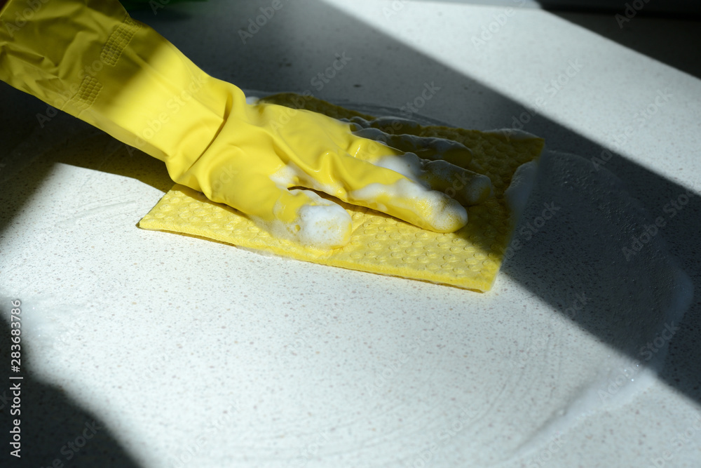 Wall mural hand in yellow rubber glove washing kitchen surface lit by the sun. home cleaning