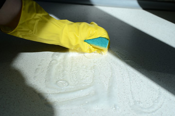 Hand in yellow rubber glove washing kitchen surface lit by the sun. Home cleaning