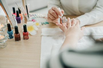 Nail care and manicure. Closeup of beautiful female hands applying transparent nail polish in home call service. woman manicurist painting lady client nails indoors on white cloth on table