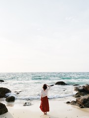 A beautiful young woman in red skirt and white shirt standing on the beach watching at blue sea...