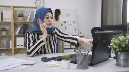Attractive arabic woman boss talking on mobile phone while using laptop at workplace sitting at desk in bright modern office. chinese colleague standing in back by shelf and white board. multi ethnic