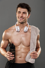 Fototapeta na wymiar Happy smiling handsome young strong sportsman isolated over grey wall background with headphones on neck holding towel and bottle with water.