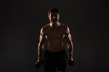 Fototapeta na wymiar Screaming handsome young strong sportsman posing isolated over black wall background holding dumbbells