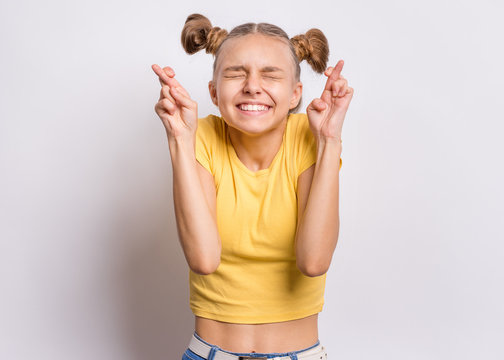 Portrait of funny teen girl crossing her fingers and wishing for good luck on gray background. Caucasian young teenager praying with crossed fingers and eyes closed. Child face expression emotions.