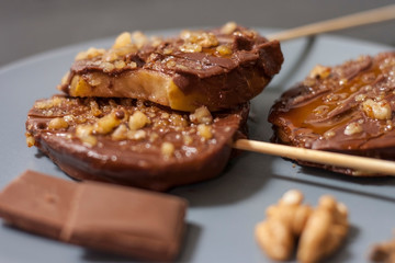 Slices of apples in chocolate, caramel glaze and walnuts on skewers on a dark background