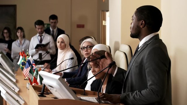 African american man giving a public talk at rostrum during international press conference while multiethnic participants sitting at table with flags of different countries and listening to speech
