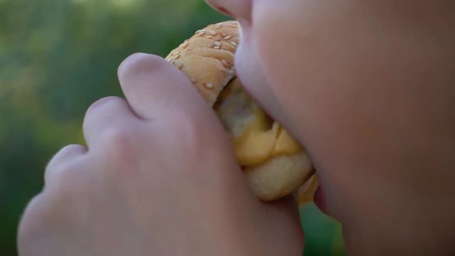 Closeup view of hungry kid eating big double cheeseburger standing outdoor. Macro of child mouth biting big meat burger. Slow motion full hd video footage.