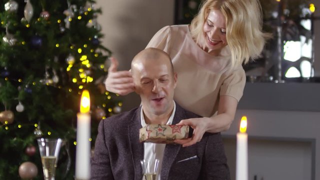 Medium shot of excited blonde 40-something Caucasian woman covering man&rsquo;s eyes and giving him New Year present, husband thanking her, both smiling and laughing, and Christmas tree in background