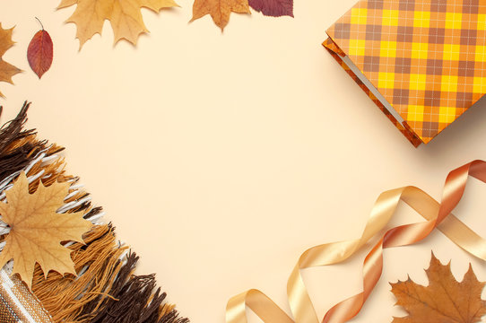 Autumn, fall concept. Checked orange gift paper bag, warm cozy blanket plaid, autumn dried leaves, holiday ribbons on pastel beige background. Flat lay top view copy space. Creative Autumn composition