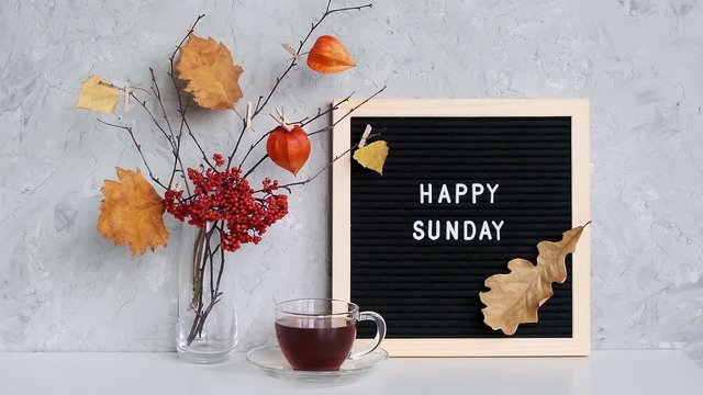 Happy Sunday text on black letter board and bouquet of branches with yellow leaves on clothespins in vase and cup of tea on table Template for postcard, greeting card Concept Hello autumn Sunday.
