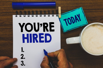 Conceptual hand writing showing You re are Hired. Business photo text New Job Employed Newbie Enlisted Accepted Recruited Man holding marker notebook clothepin reminder wooden table coffee