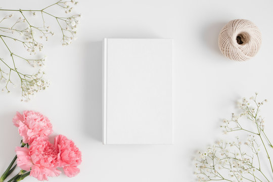 Top view of a book mockup with a bouquet of pink carnations, workspace accessories and a gypsophila  on a white table.