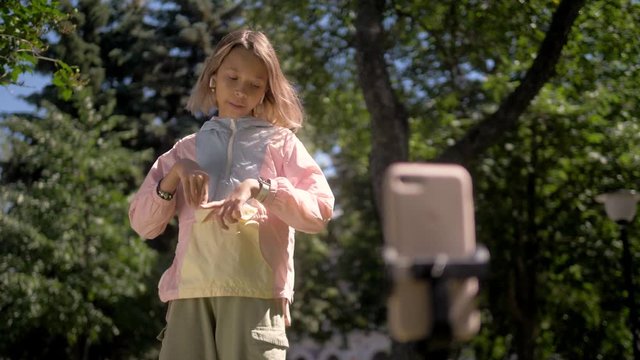 School girl take a selfie on a phone on a sunny day with tripod selfie stick, kid dance and sign in front of camera in park, use mask application film maker