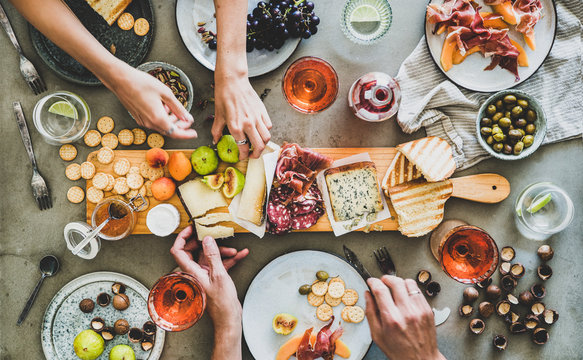 Mid-summer picnic with wine and snacks. Flat-lay of charcuterie and cheese board, rose wine, nuts, olives and peoples hands over concrete table background, top view. Family, friends holiday gathering