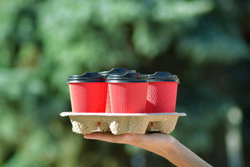 Four red paper glasses for coffee with black covers on a stand in hand on a blurred background of green trees