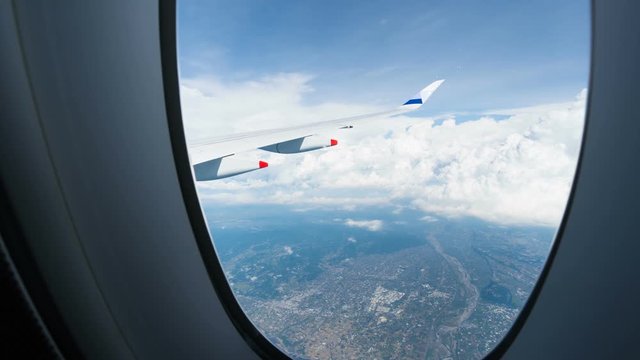 Timelapse view through plane illuminator of passenger jet wing flying above blue sea, view of light white clouds and bright blue sky