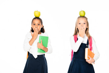 two shocked multicultural schoolgirls with apples on heads covering mouths with hands isolated on white