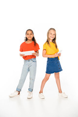 Obraz premium full length view of two adorable multicultural friends holding smartphone and digital tablet on white background