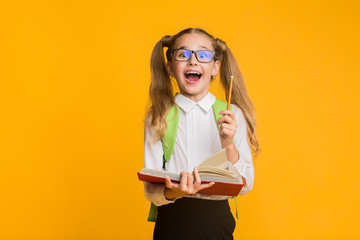 Excited Elementary Schoolgirl Holding Book And Pencil On Yellow Background