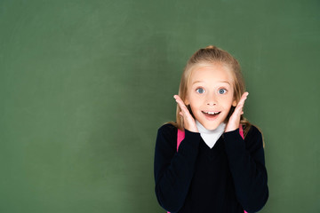 excited schoolgirl smiling at camera while standing near green green chalkboard