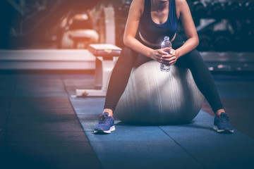 Fototapeta premium Asian beautiful girls wear sports bra She sat on a yoga ball inside the gym. After exercising. Relaxed after exercising and drinking water. Bottled water in hand. The concept of lifestyle and healthy.