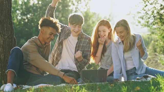 Group of cheerful multiethnic students having fun together and using laptop computer while sitting outdoors in park