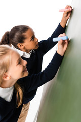 two adorable multicultural schoolgirls writing on green chalkboard