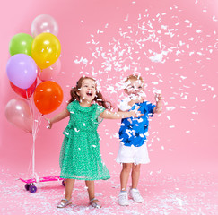 Сute little girl and boy joyfully throws confetti isolated on pink background. Celebrating brightful  birthday party with colorful balloons. expressing positivity