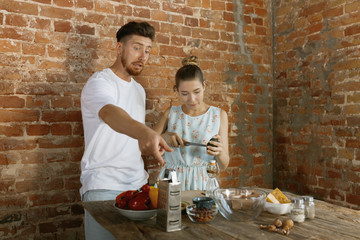 Fototapeta na wymiar Young caucasian happy couple cooking together using vegetables, cheese, eggs and nuts in recipe against brick wall in their kitchen. Nutrition, healthy food, family, relations, domestic life concept.