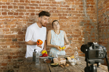 Fototapeta na wymiar Vlogger and blogger freelance job, food concept. Young caucasian couple cooking together and recording live video for vlog and social media with professional camera against brick wall in their kitchen