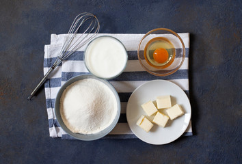 Ingredients for crepes and pancakes on a dark background. Flour, egg, milk, butter, whisk. Top view. Space for text