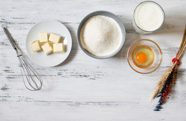 Ingredients for crepes and pancakes on a white wooden background. Flour, egg, milk, butter, whisk. Top view. Space for text.