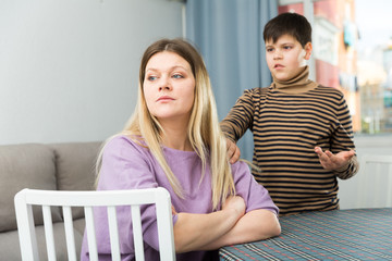 Upset mother sitting at table after arguing with son indoors
