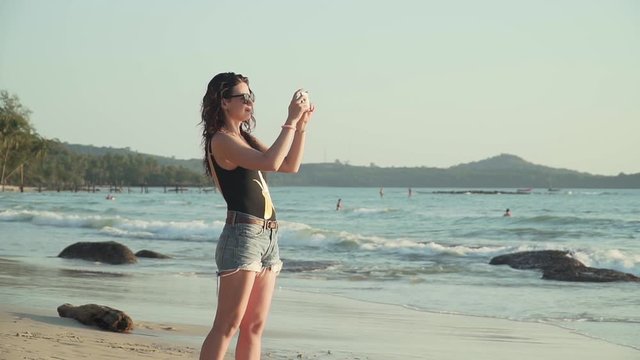 Pretty young caucasian woman in sunglasses and swimsuit takes pictures of the beautiful sunset over sea on tropical island beach. Happy girl on vacation photographs on smartphone. Slow motion footage