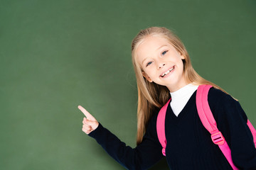 smiling schoolgirl pointing with finger at green chalkboard while smiling at camera