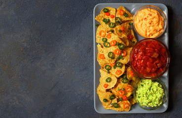 Mexican yellow corn tortilla chips nachos with jalapeno, guacamole, cheese sauce and salsa on dark blue background