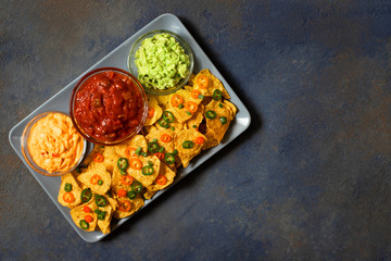 Mexican yellow corn tortilla chips nachos with jalapeno, guacamole, cheese sauce and salsa on dark blue background
