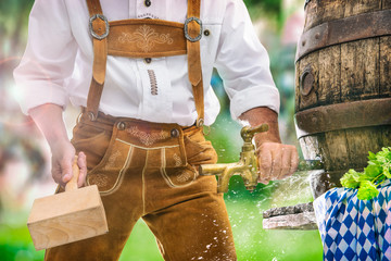 Bavarian man in leather trousers taps a wooden barrel of beer in the garden