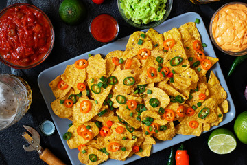 Mexican yellow corn tortilla chips nachos with jalapeno, guacamole, cheese sauce and salsa on black background. Glasses with beer, lime, chili peppers, cherry tomatoes