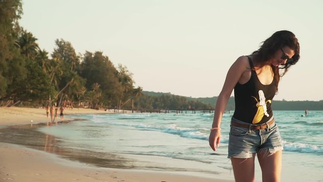 Young happy caucasian woman is spinning and dancing on beach of tropical island at sunset. Smiling pretty girl in sunglasses enjoys freedom and vacation on sandy coast by sea. Slow motion footage.