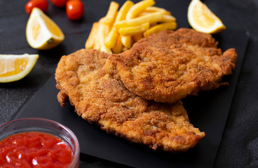 Traditional chicken schnitzel served with french fries, sauce and lemon. Black background. Close-up view