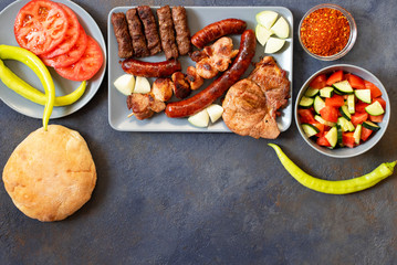Traditional Serbian and Balkan grilled meat called mesano meso. Balkan barbeque (rostilj) served with Serbian salad, hot peppers, bread, tomato, onions, and paprika powder. Dark background. Top view