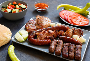 Traditional Serbian and Balkan grilled meat called mesano meso. Balkan barbeque (rostilj) served with Serbian salad, hot peppers, bread, tomato, onions, and paprika powder. Dark background. Close-up