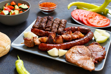 Traditional Serbian and Balkan grilled meat called mesano meso. Balkan barbeque (rostilj) served with Serbian salad, hot peppers, bread, tomato, onions, and paprika powder. Dark background. Close-up