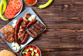 Traditional Serbian and Balkan grilled meat called mesano meso. Balkan barbeque (rostilj) served with Serbian salad, hot peppers, bread, tomato, onions, and paprika powder. Wooden background. Top view