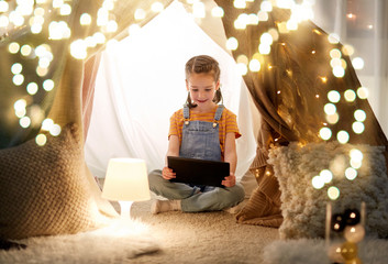 childhood, technology and hygge concept - happy little girl with tablet pc computer in kids tent at home