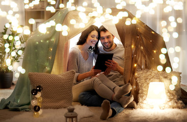 leisure, hygge, technology and people concept - happy couple with tablet pc computer in kids tent at home