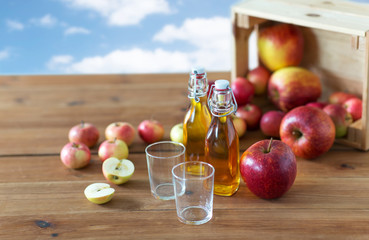 fruits, food and harvest concept - two glasses and bottles of apple juice or cider on wooden table over blue sky and clouds background