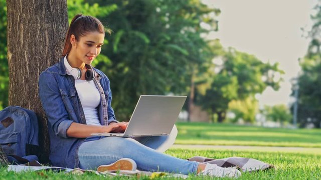 Happy brunette woman using laptop computer while sitting near the tree on grass in park